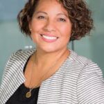 A confident woman with curly hair smiling in an office environment, conveying professionalism and approachability.